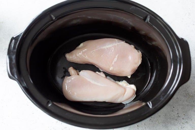 Two raw chicken breasts placed inside a black slow cooker on a light-colored surface.