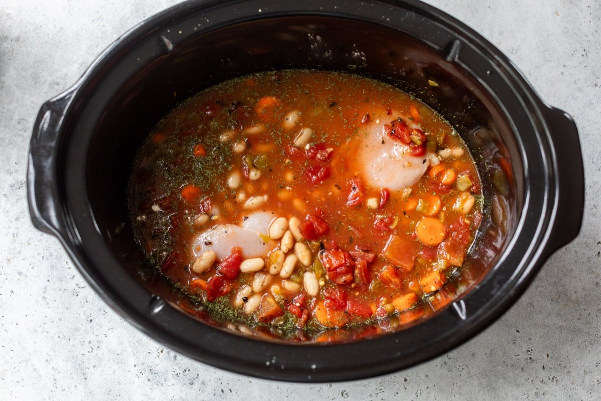 Raw chicken, beans, tomatoes, and vegetables in broth inside a black slow cooker, ready to be cooked.