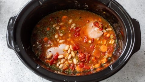 Raw chicken, beans, tomatoes, and vegetables in broth inside a black slow cooker, ready to be cooked.