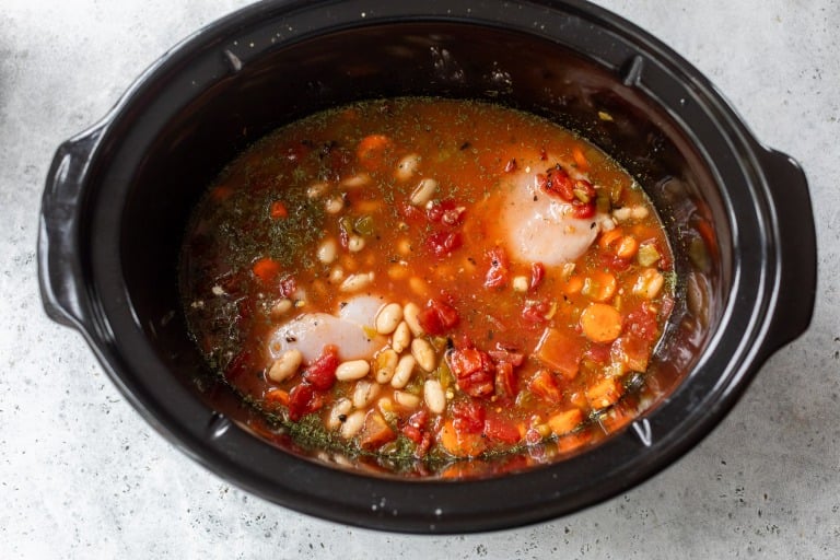 Raw chicken, beans, tomatoes, and vegetables in broth inside a black slow cooker, ready to be cooked.
