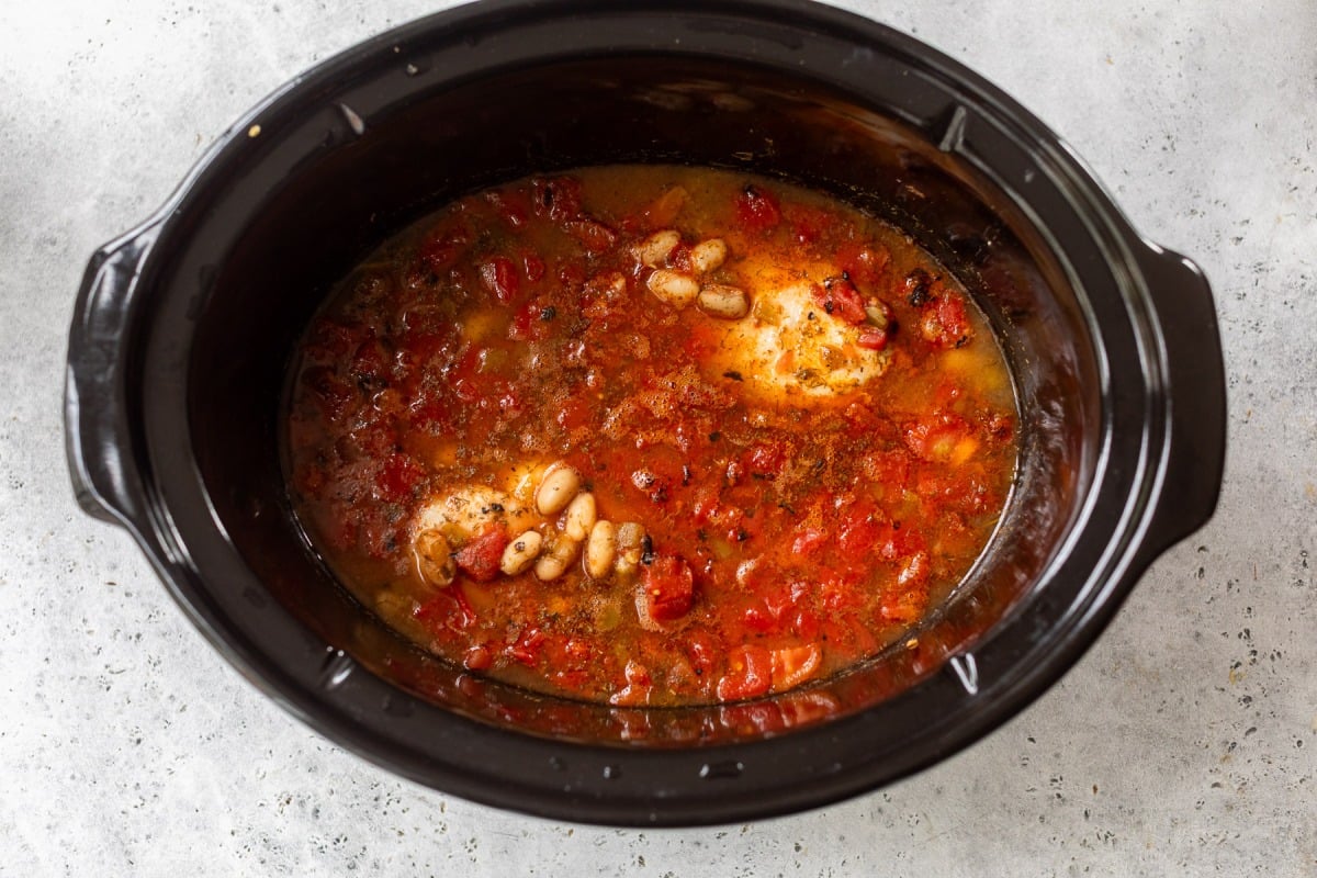 Chicken breasts, beans, diced tomatoes, and spices in a slow cooker with broth, ready to be cooked.