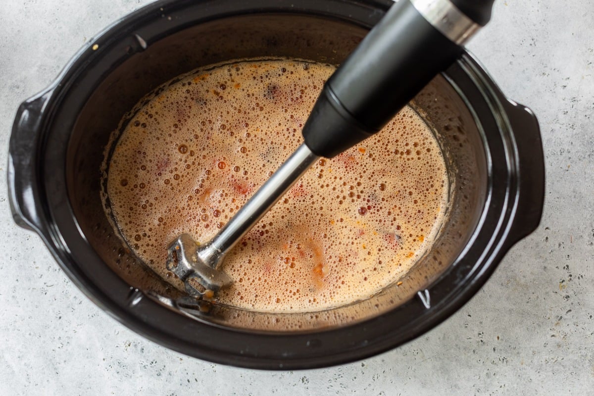 An immersion blender blending chili inside a black slow cooker on a light gray countertop.