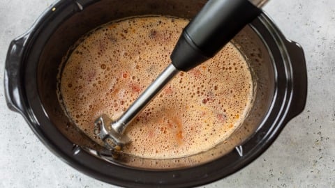 An immersion blender blending chili inside a black slow cooker on a light gray countertop.