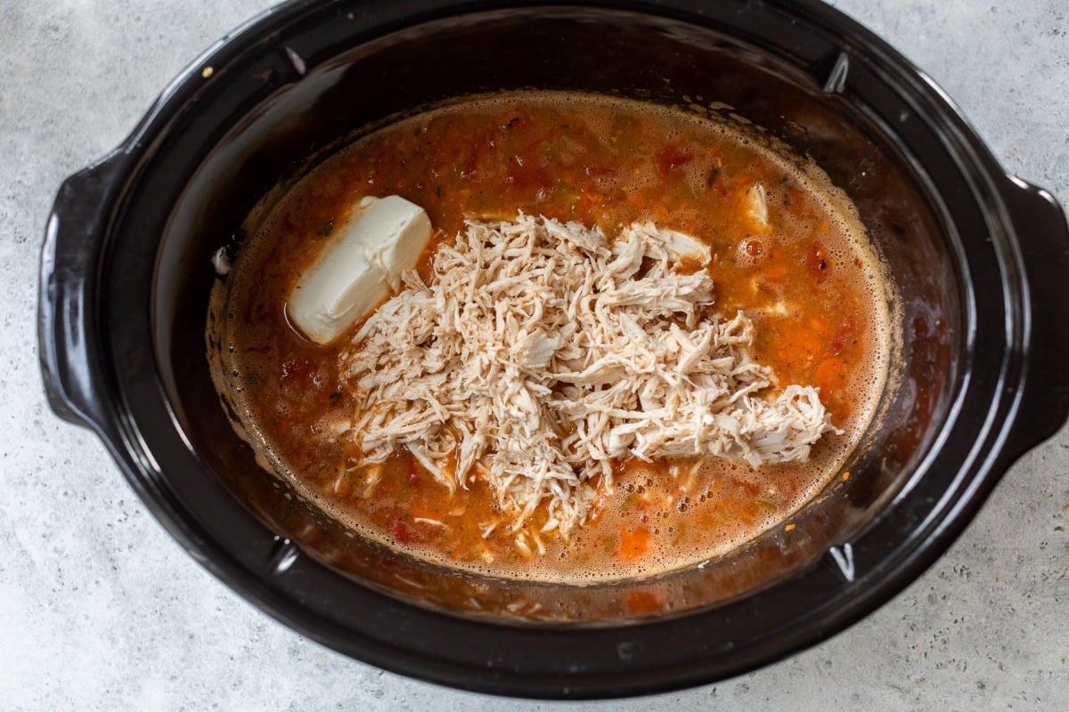 Shredded chicken, broth, diced tomatoes, and a block of cream cheese in a black slow cooker.
