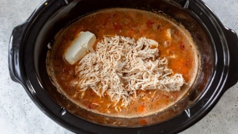 Shredded chicken, broth, diced tomatoes, and a block of cream cheese in a black slow cooker.