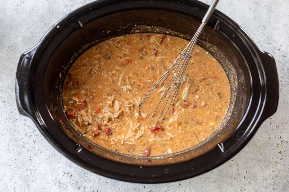 A black slow cooker filled with a creamy, mixed soup and a metal whisk resting inside.
