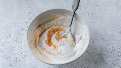 A bowl of yogurt and broth being mixed with a spoon, showing swirls of white and orange on a light gray surface.
