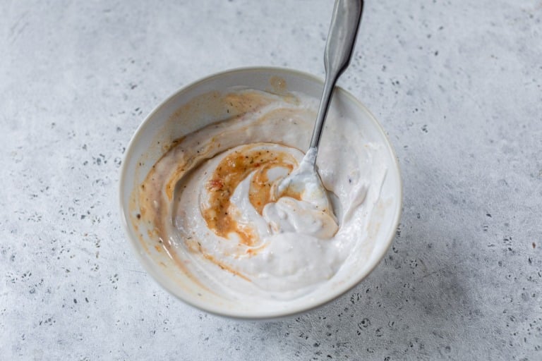 A bowl of yogurt and broth being mixed with a spoon, showing swirls of white and orange on a light gray surface.
