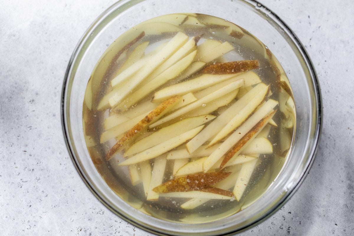 Thin potato slices soaking in a glass bowl filled with water, on a light gray surface.