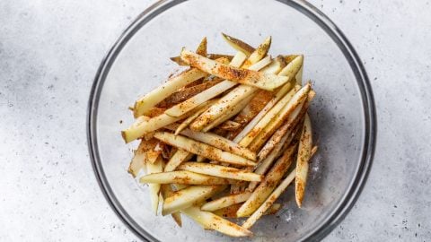Raw potato fries in a glass bowl, sprinkled with spices, on a light gray surface.