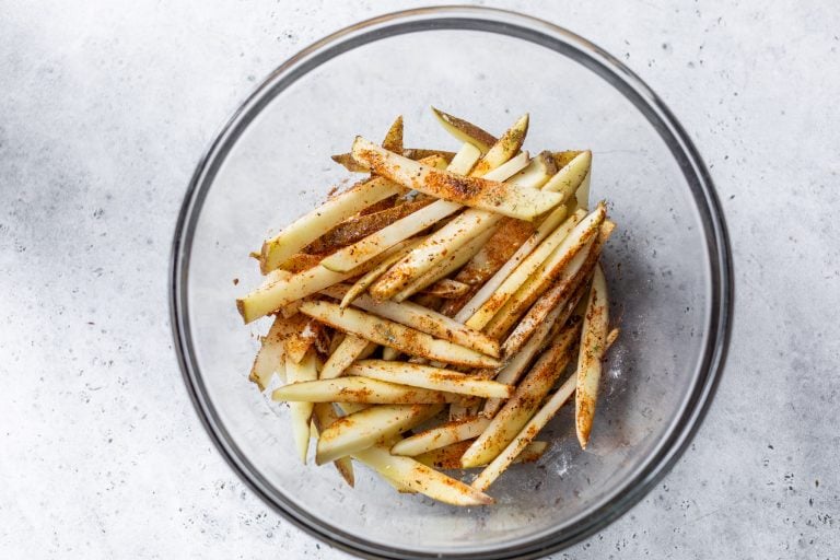Raw potato fries in a glass bowl, sprinkled with spices, on a light gray surface.