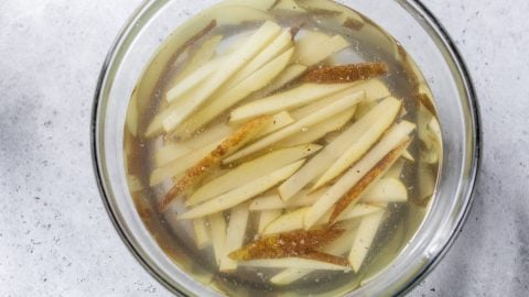 Thin potato slices soaking in a glass bowl filled with water, on a light gray surface.