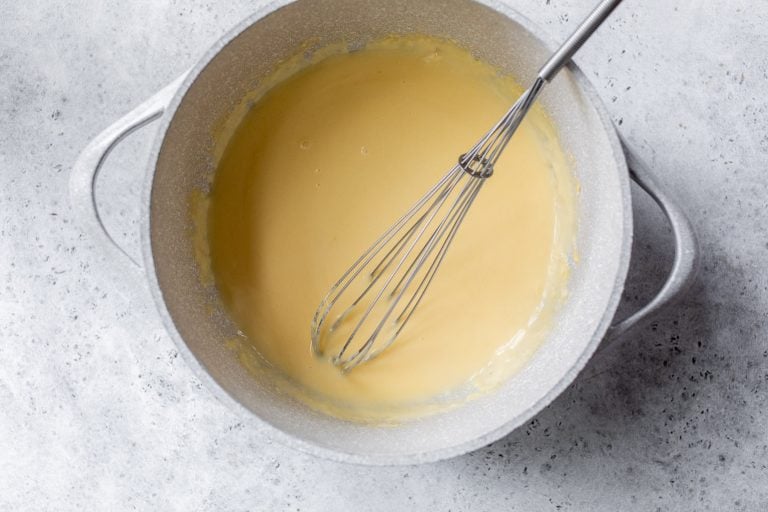 A metal whisk stirring creamy cheese sauce in a light gray pot on a textured gray surface.