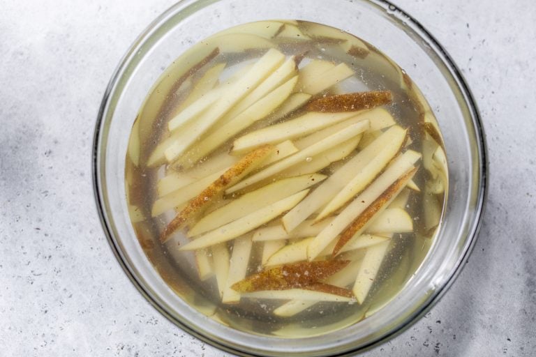 Thin potato slices soaking in a glass bowl filled with water, on a light gray surface.