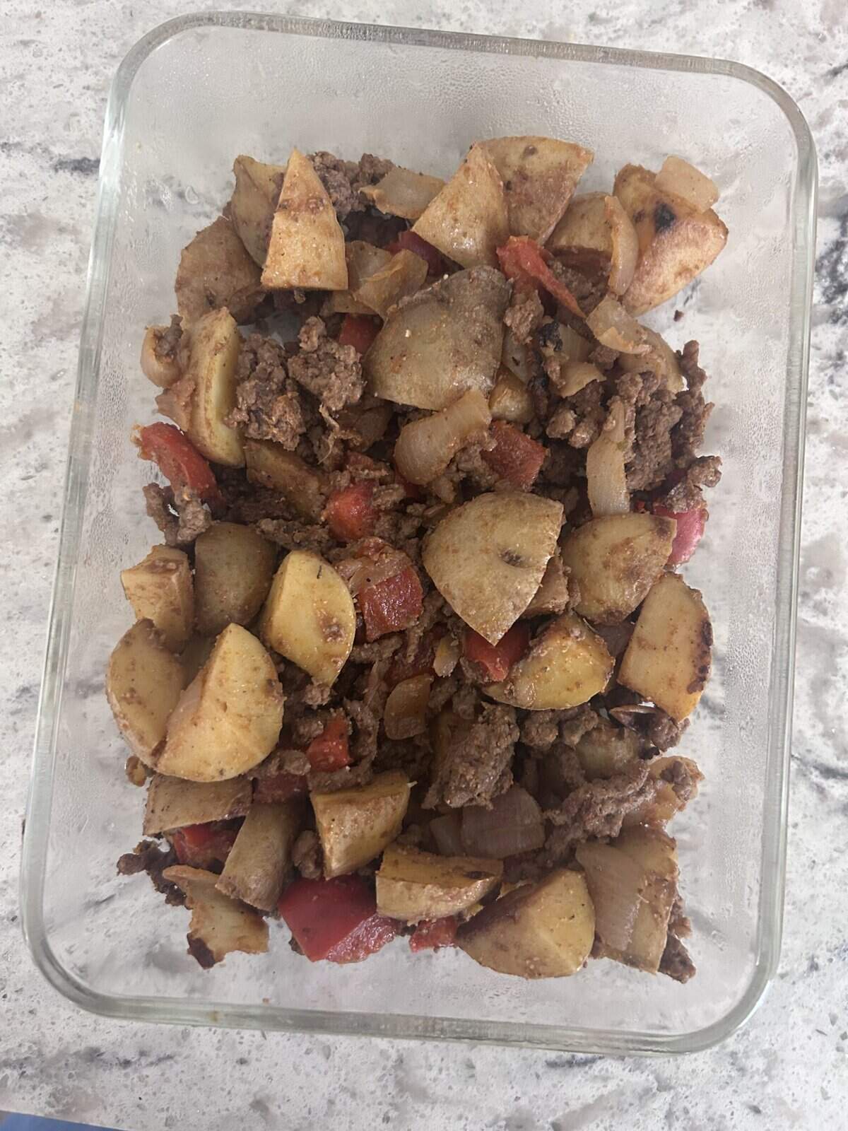 Glass dish with a cooked mixture of ground beef, diced potatoes, onions, and red bell peppers on a light countertop.