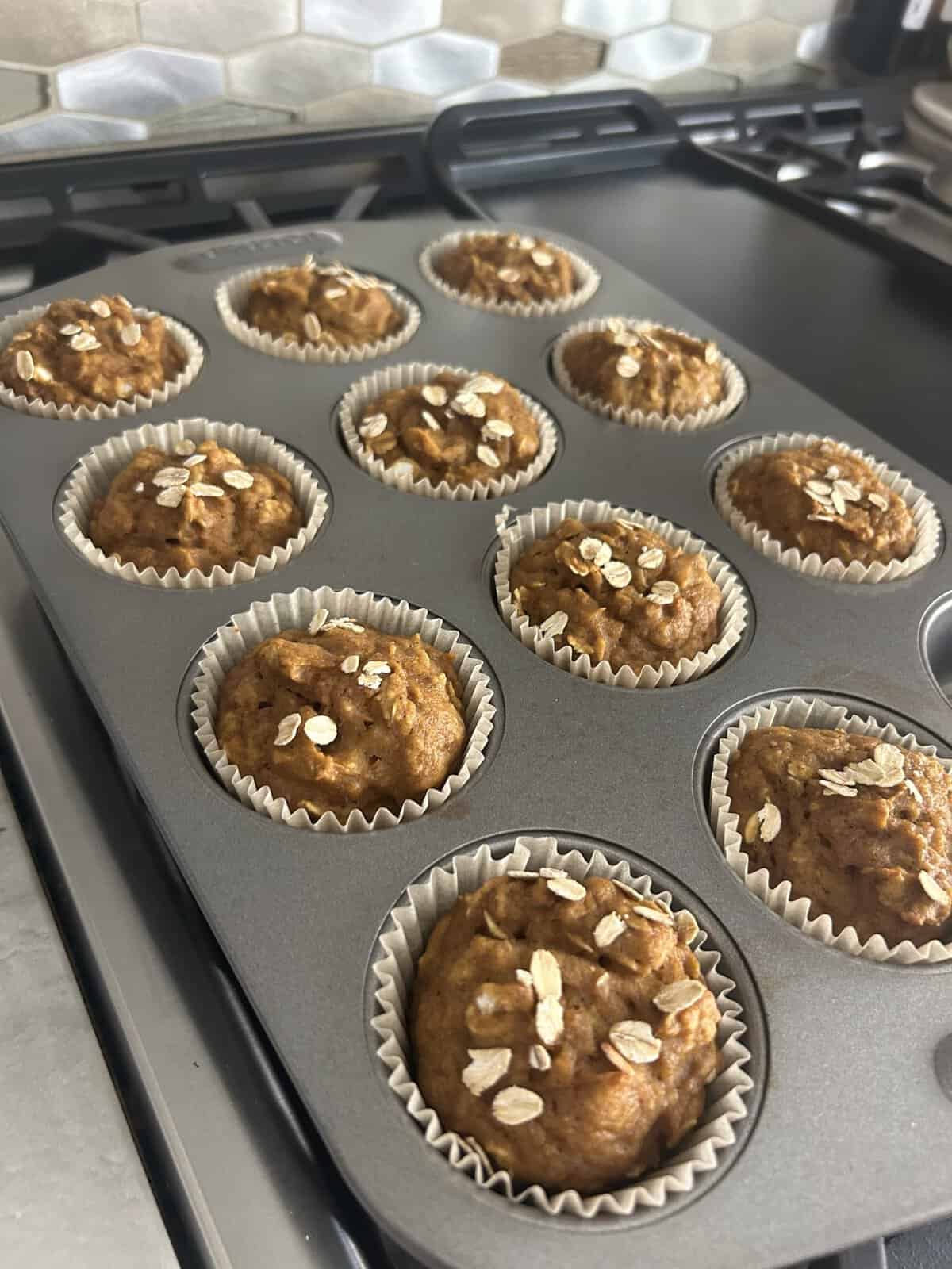 A muffin tray with ten baked muffins topped with oats, sitting on a stovetop in a kitchen.