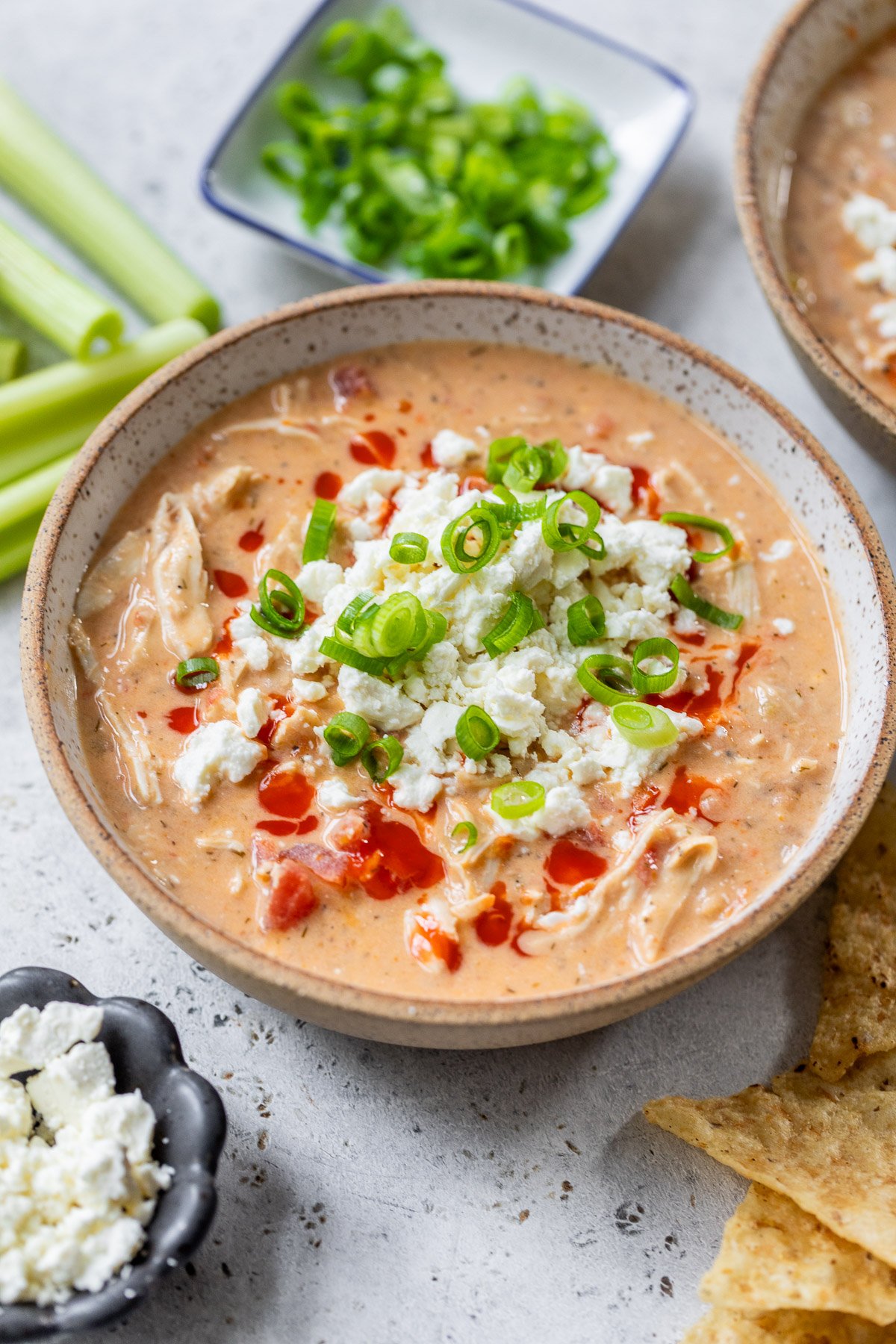 Bowl of creamy Buffalo chicken chili topped with crumbled cheese, green onions, and hot sauce, with chips and veggies nearby.