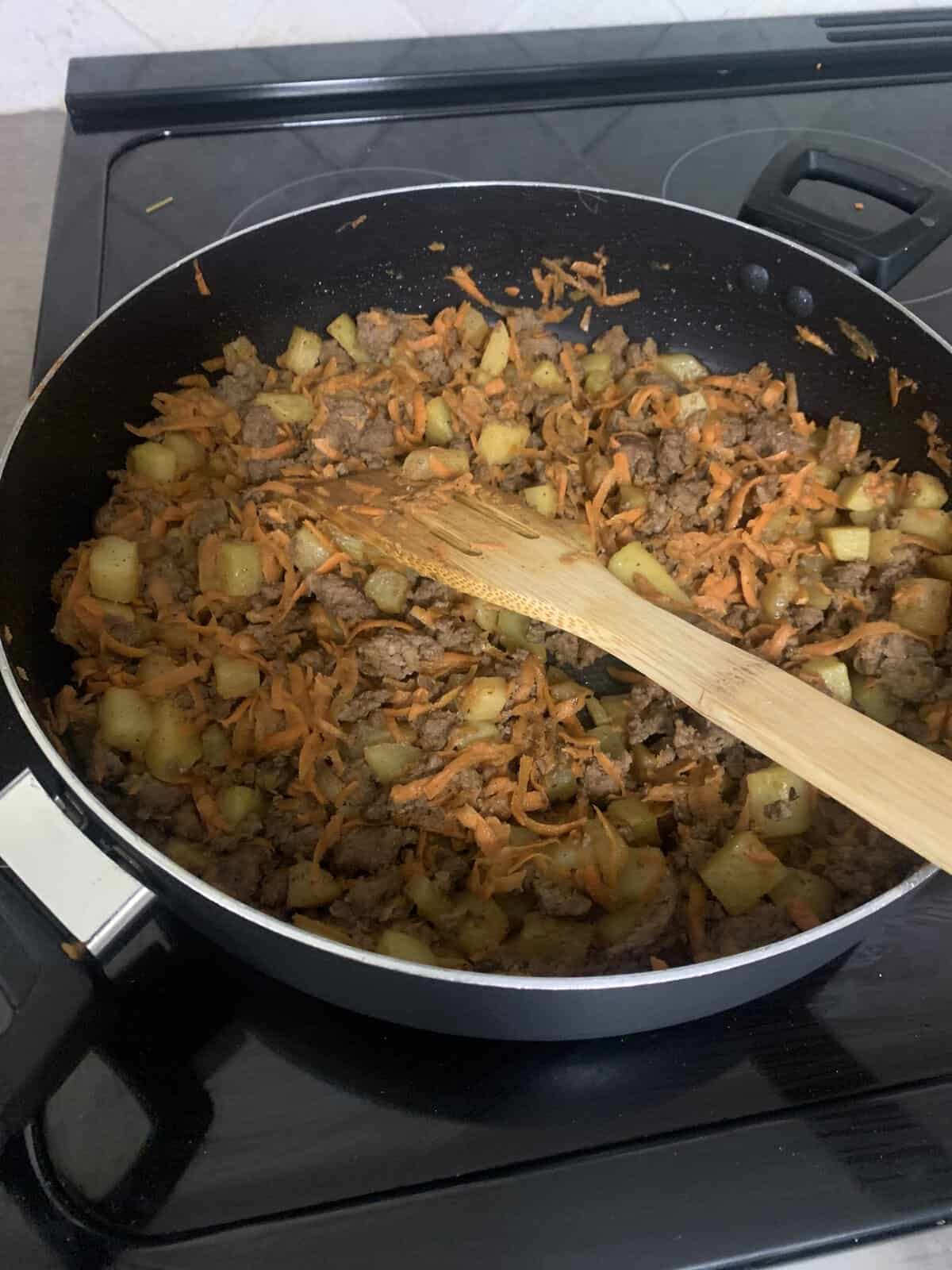 A pan on a stove with cooked ground beef, diced potatoes, and shredded carrots, stirred with a wooden spatula.