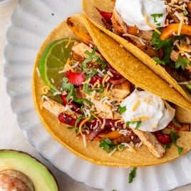 Two sheet pan chicken fajita tacos with peppers, cheese, sour cream, and cilantro on a plate, with lime and half an avocado nearby.