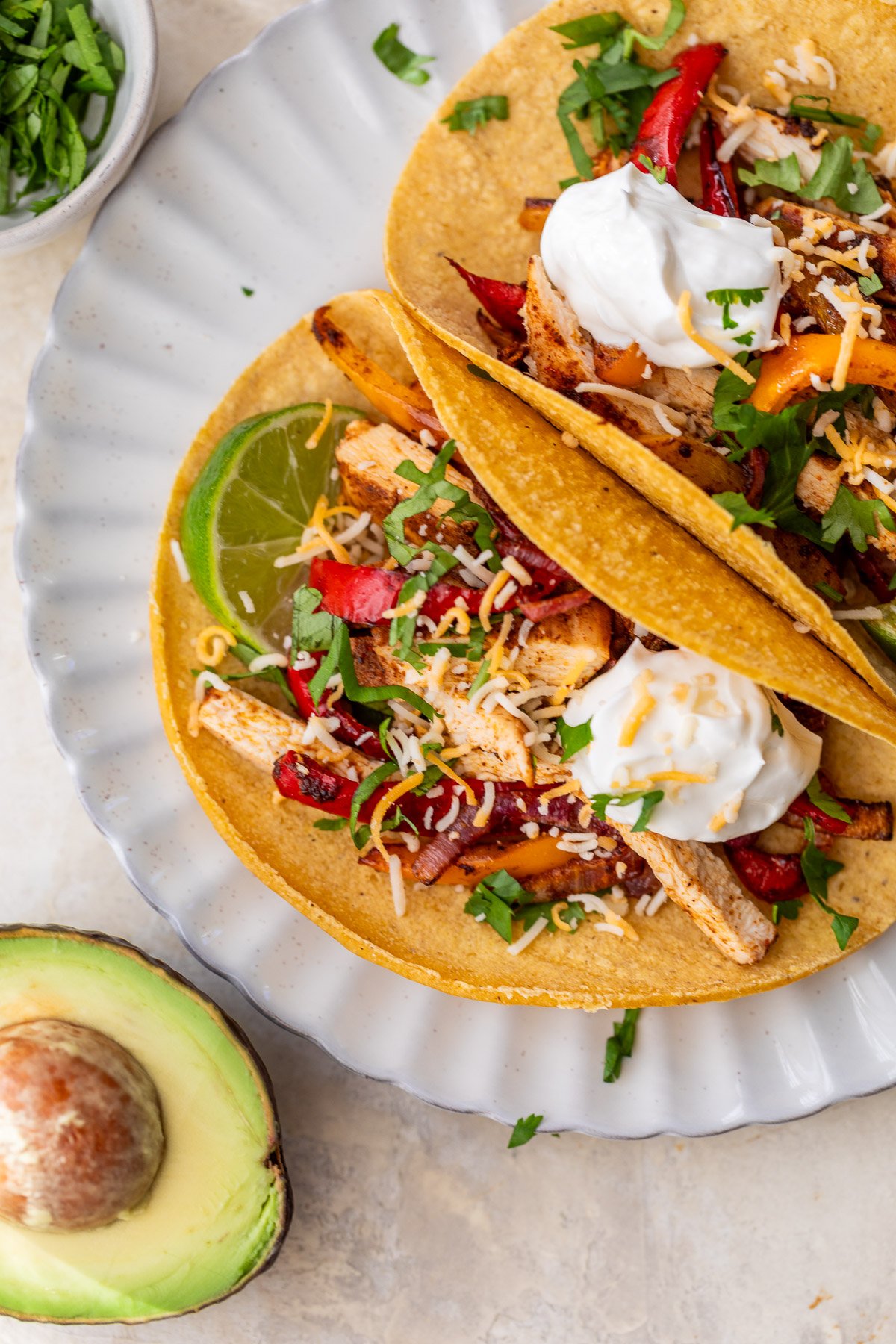 Two sheet pan chicken fajita tacos with peppers, cheese, sour cream, and cilantro on a plate, with lime and half an avocado nearby.