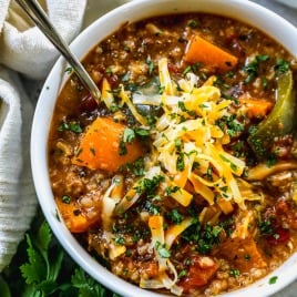 A bowl of stuffed pepper soup topped with shredded cheese and chopped herbs, with a spoon and fresh cilantro nearby.