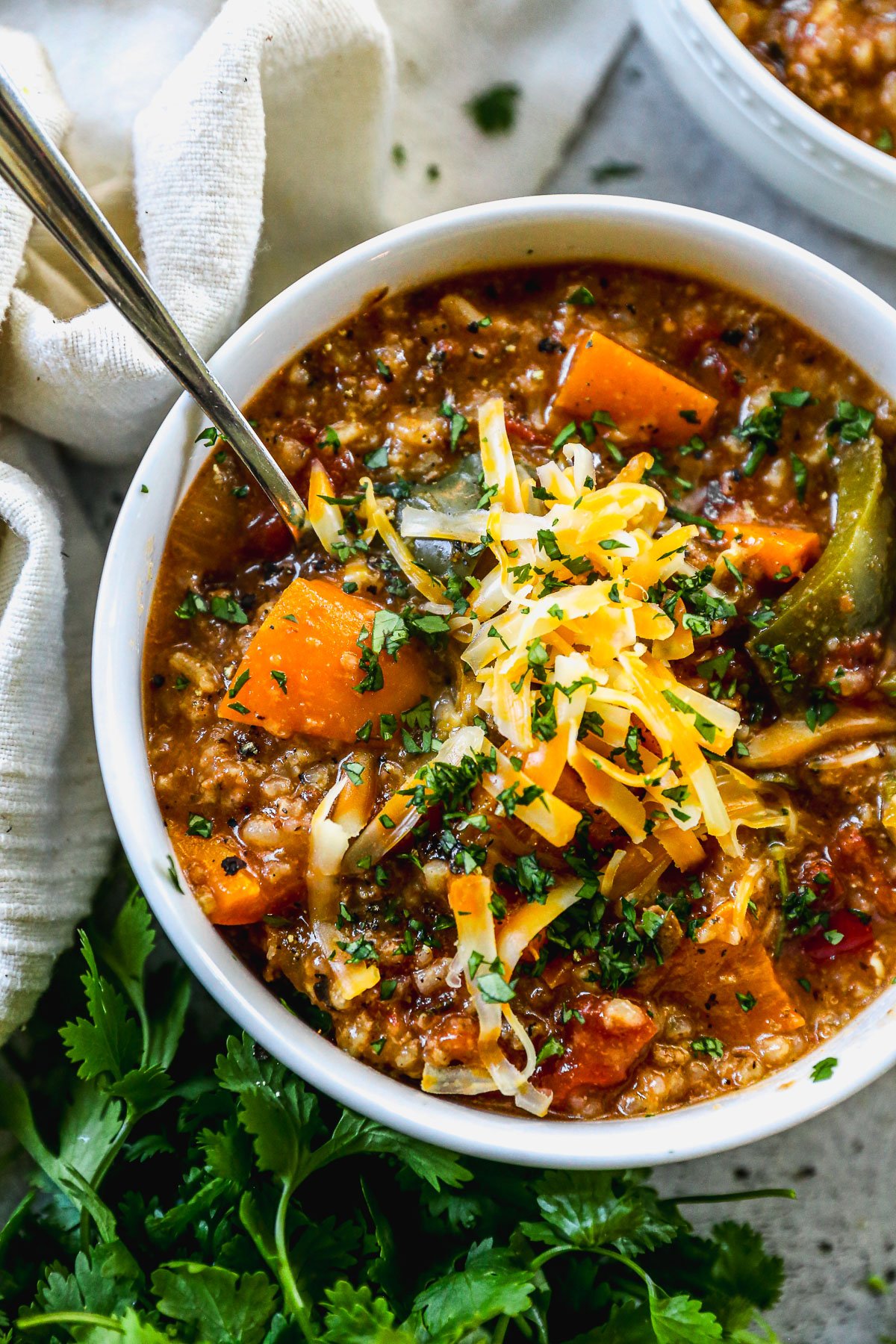 A bowl of stuffed pepper soup topped with shredded cheese and chopped herbs, with a spoon and fresh cilantro nearby.