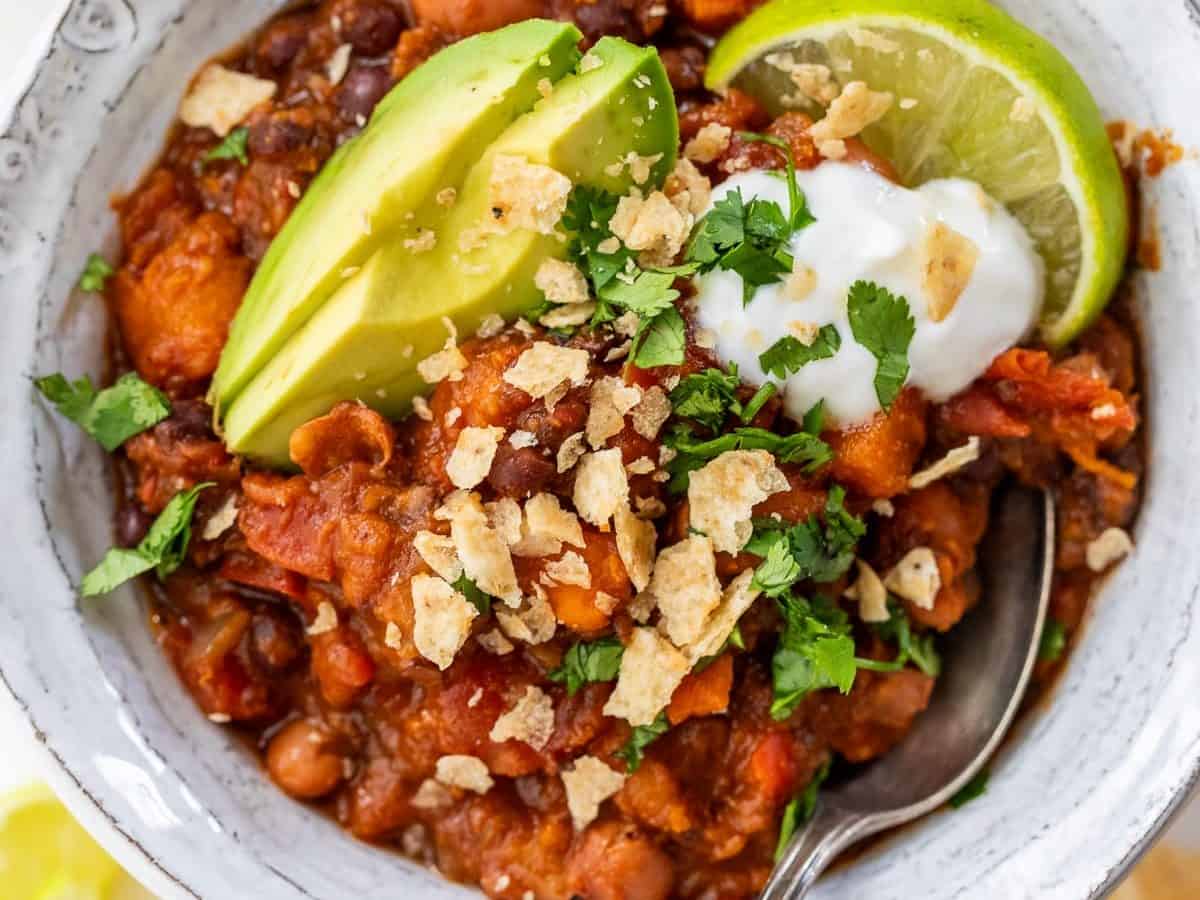 Vegan chili in bowl with sliced avocado, lime wedge, sour cream, crushed tortilla chips, and cilantro.