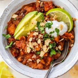 Vegan chili in bowl with sliced avocado, lime wedge, sour cream, crushed tortilla chips, and cilantro.