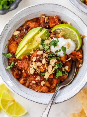 Vegan chili in bowl with sliced avocado, lime wedge, sour cream, crushed tortilla chips, and cilantro.