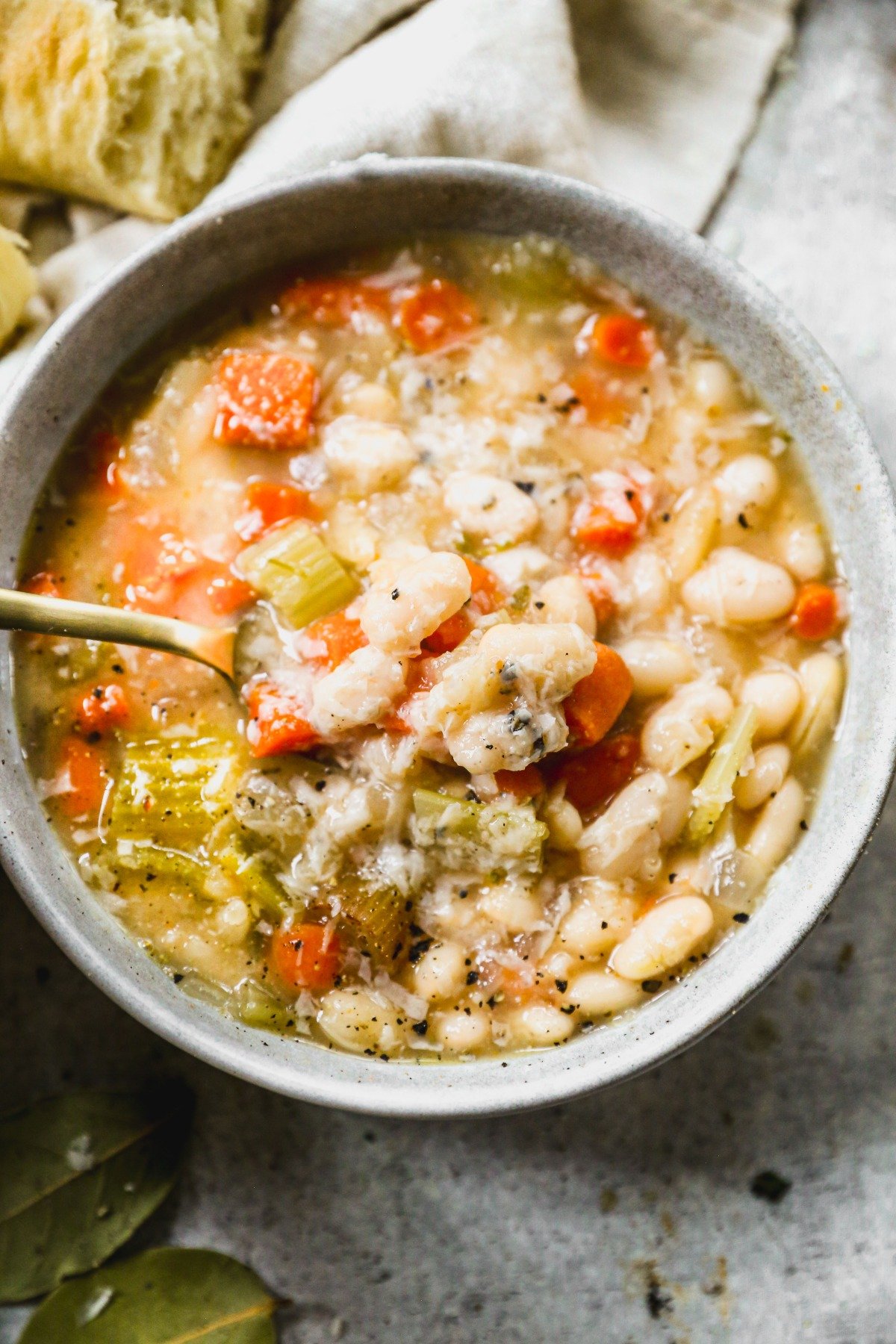 A bowl of white bean soup with carrots and celery, topped with herbs, and a spoon inside the bowl.