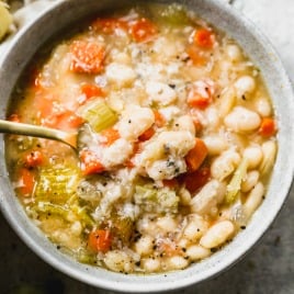 A bowl of white bean soup with carrots and celery, topped with herbs, and a spoon inside the bowl.