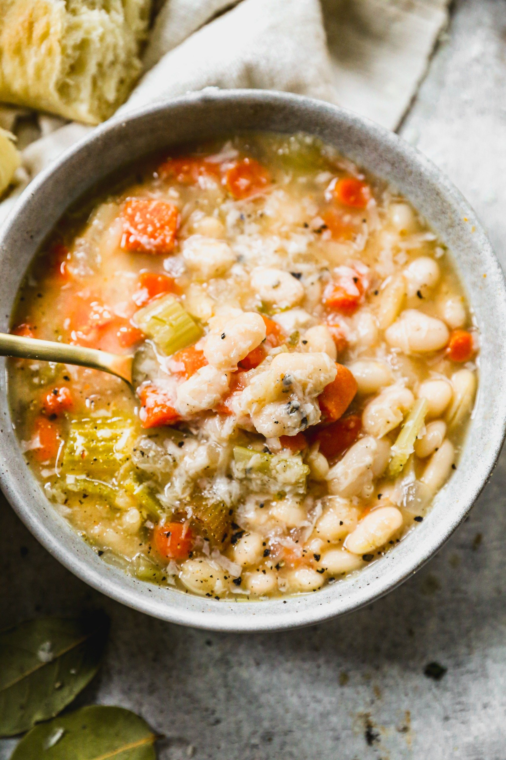 A bowl of white bean soup with carrots and celery, topped with herbs, and a spoon inside the bowl.
