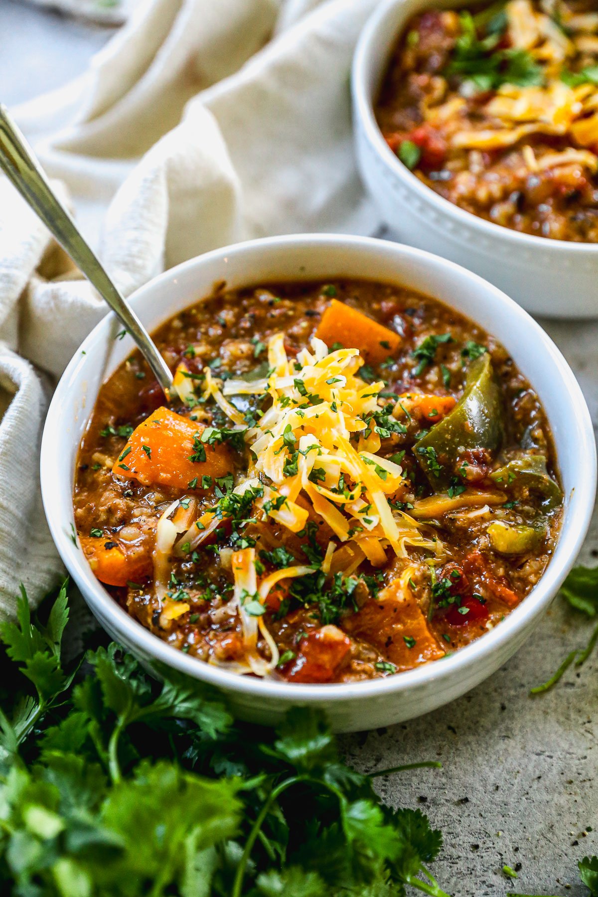 A bowl of hearty stuffed pepper soup topped with shredded cheese and chopped herbs, with a spoon and fresh cilantro nearby.