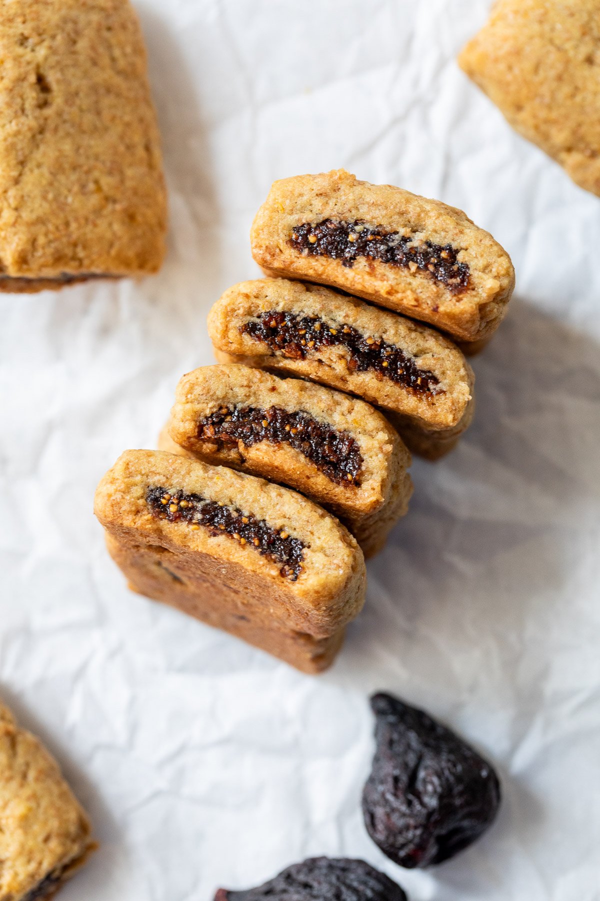 Four fig bars, one cut open to show the fig filling, on crumpled white paper with dried figs nearby.