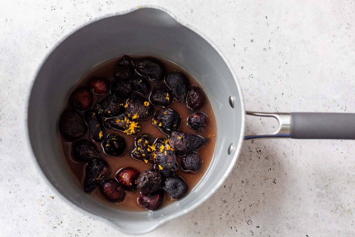 A saucepan with dried figs simmering in liquid, garnished with orange zest on a light gray surface.