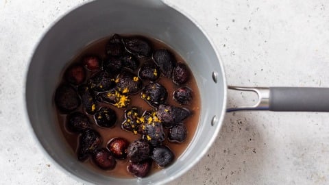 A saucepan with dried figs simmering in liquid, garnished with orange zest on a light gray surface.