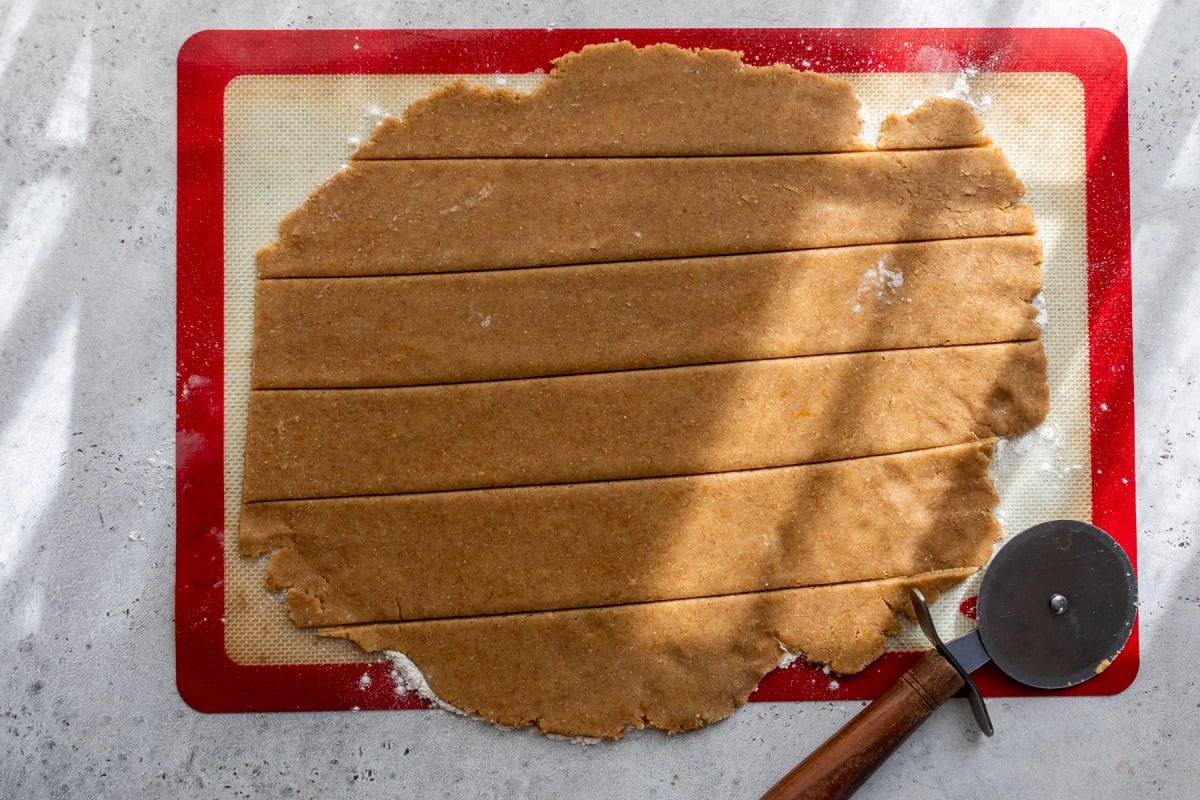 Rolled out fig bar dough on a red-edged baking mat, cut into strips with a pizza cutter resting nearby.