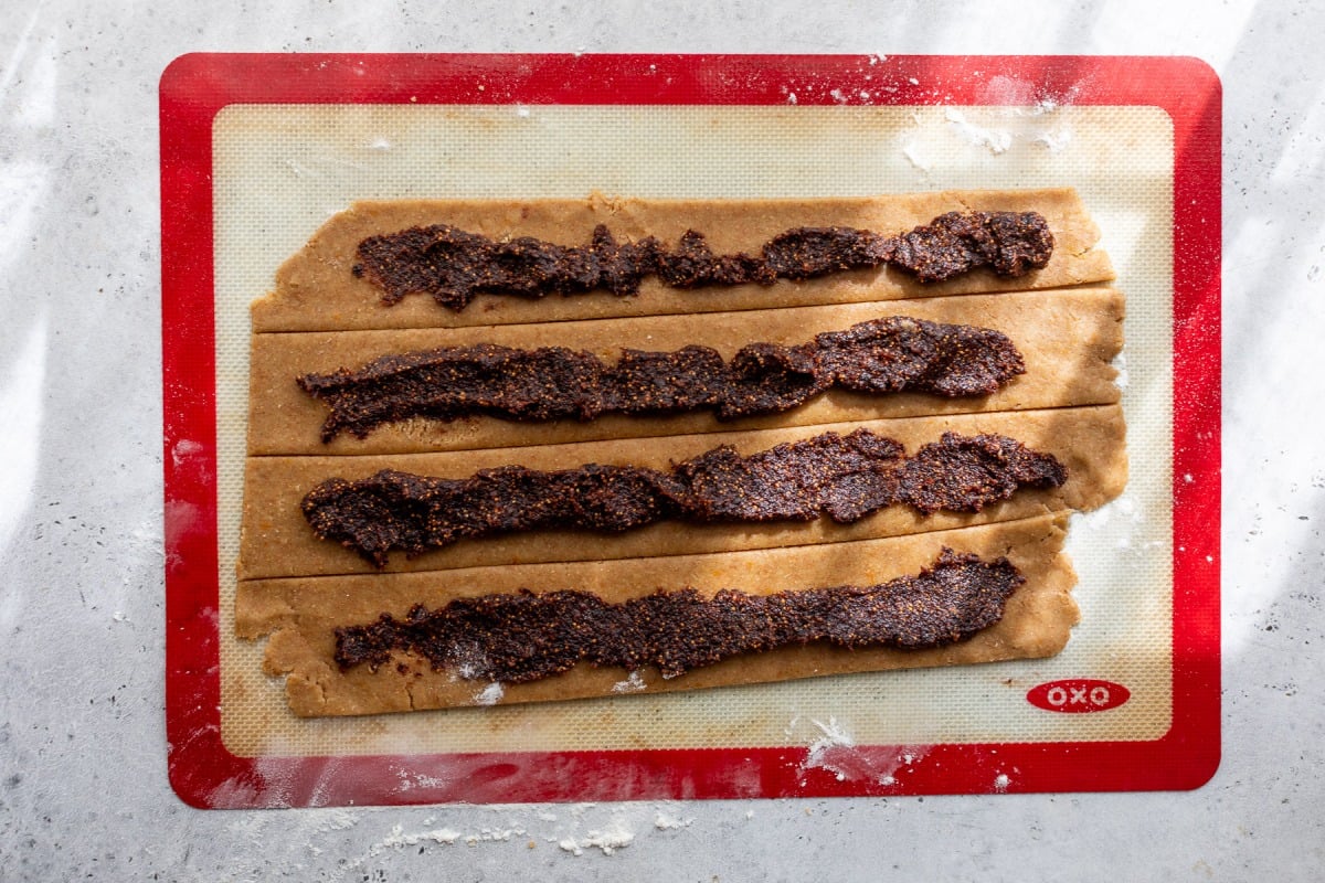 Dough strips on a silicone mat topped with a thick, dark fig filling, ready to be rolled or folded.