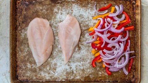 Raw chicken breasts and sliced red onion, red bell pepper, and yellow bell pepper on a baking sheet.