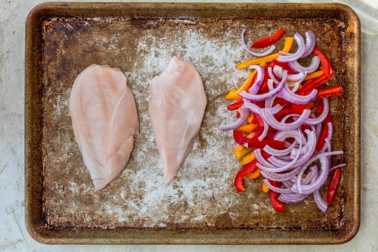 Raw chicken breasts and sliced red onion, red bell pepper, and yellow bell pepper on a baking sheet.