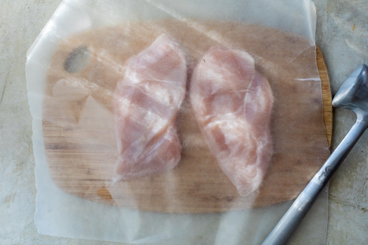 Two raw chicken breasts on a wooden board covered with parchment paper, meat mallet placed to the right.