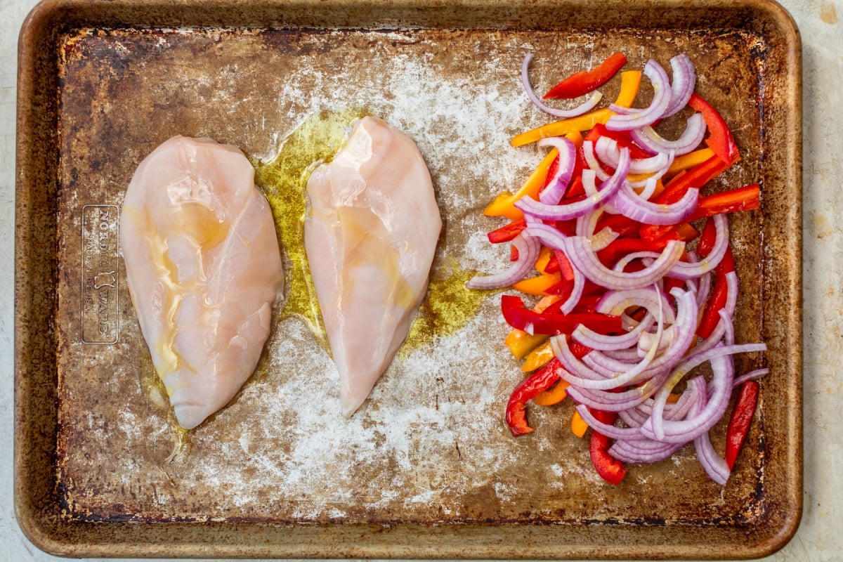 Two raw chicken breasts and sliced red onions and bell peppers on a baking sheet, drizzled with olive oil.