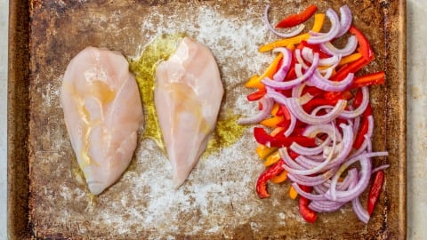 Two raw chicken breasts and sliced red onions and bell peppers on a baking sheet, drizzled with olive oil.