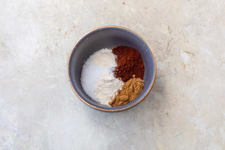 A bowl containing four different spices and seasonings for chicken fajitas on a light, textured surface.