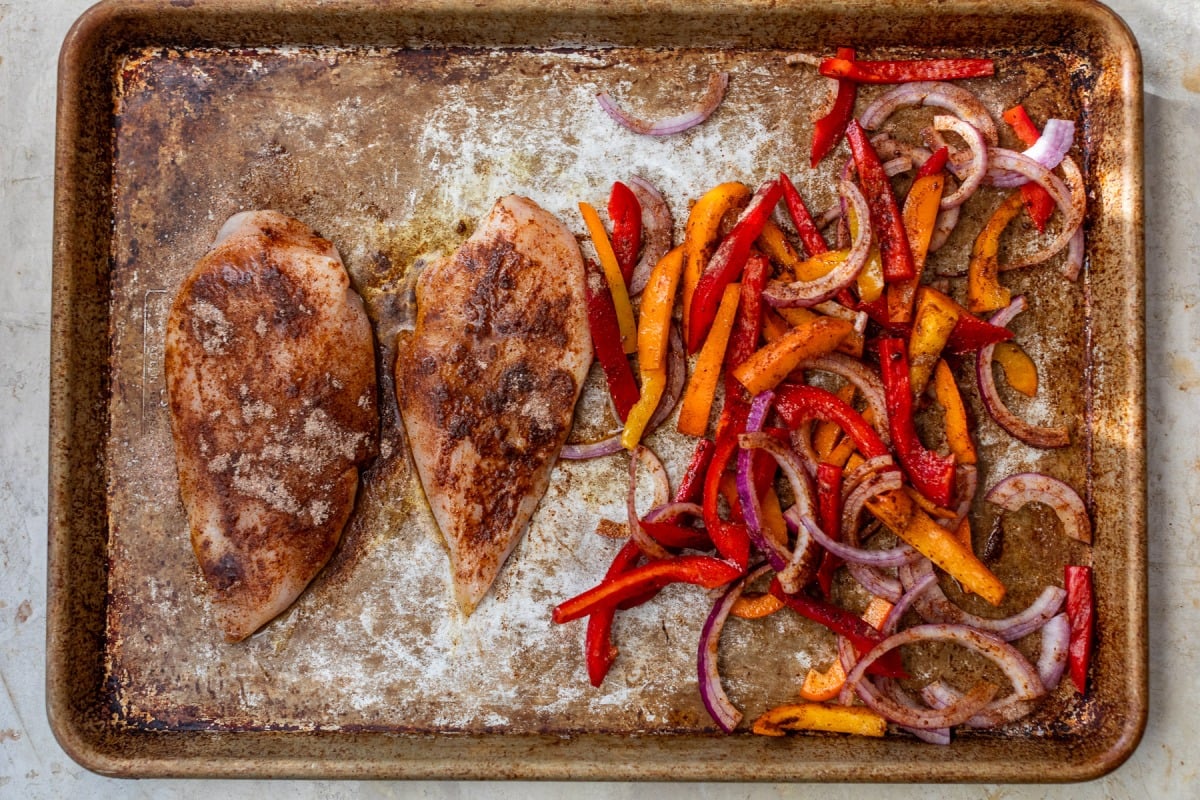 Two seasoned chicken breasts and sliced red onions and bell peppers on a worn sheet pan for fajitas.
