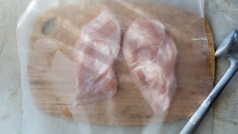 Two raw chicken breasts on a wooden board covered with parchment paper, meat mallet placed to the right.