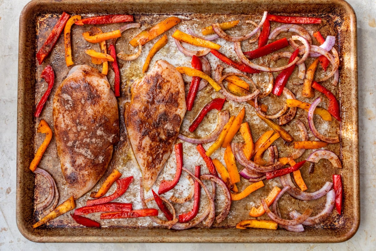 Sheet pan chicken fajitas with two seasoned chicken breasts, sliced red and yellow bell peppers, and red onion, all roasted.