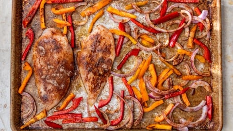 Sheet pan chicken fajitas with two seasoned chicken breasts, sliced red and yellow bell peppers, and red onion, all roasted.
