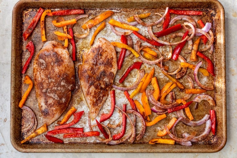 Sheet pan chicken fajitas with two seasoned chicken breasts, sliced red and yellow bell peppers, and red onion, all roasted.