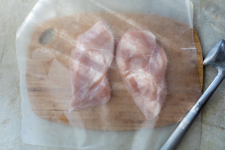 Two raw chicken breasts on a wooden board covered with parchment paper, meat mallet placed to the right.